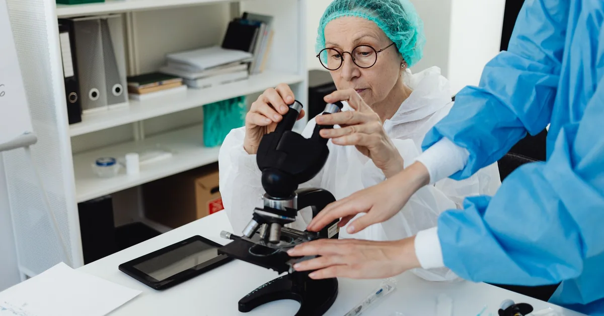 Senior researcher working with a microscope in a laboratory setting, assisted by a colleague.