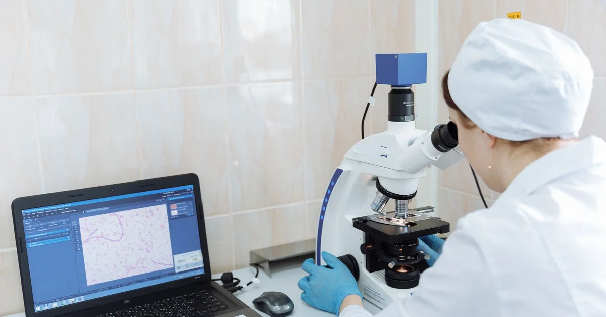 Scientist in lab coat using microscope and laptop in a laboratory setting.