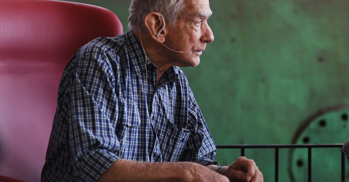 A senior man speaks into a headset microphone during an indoor event.