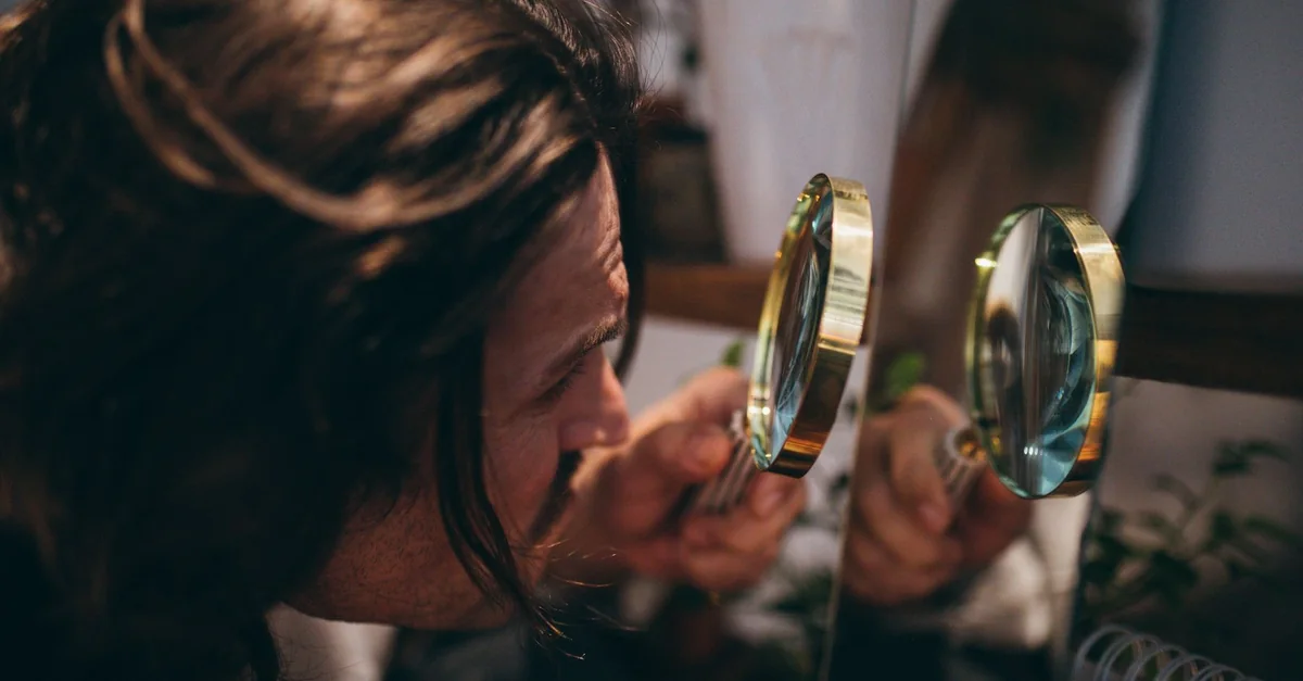 Close-up shot of a man with long hair using a magnifying glass to inspect his reflection in a mirror.