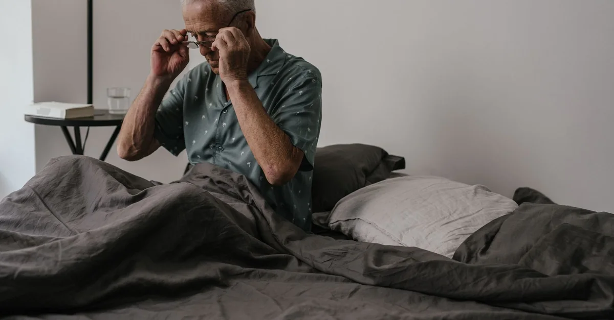Senior man adjusts glasses while sitting in bed, expressing morning routine and daily life.
