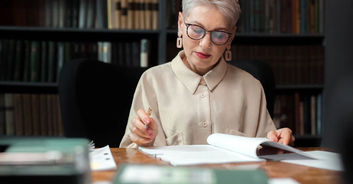 A senior woman wearing eyeglasses and a beige top reading documents in a library.