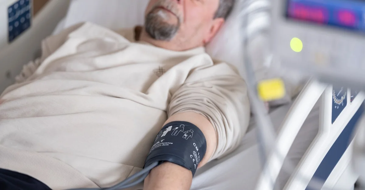 Elderly man resting in hospital bed with blood pressure monitor attached, symbolizing healthcare and medical care.