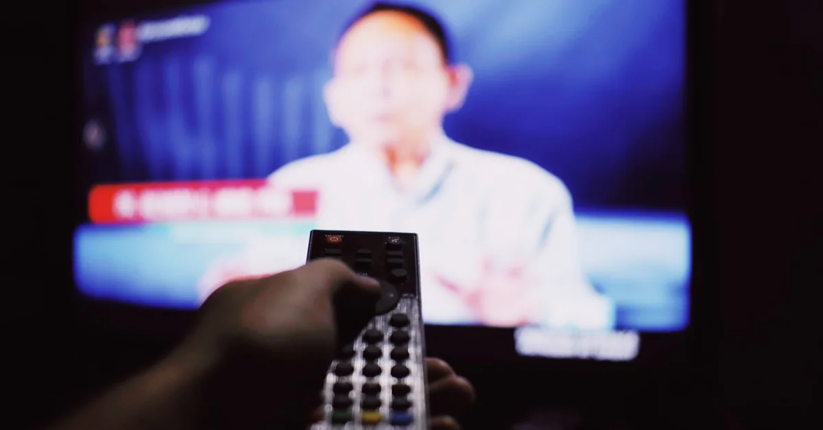 Crop anonymous person with remote controller switching channels while watching TV on blurred background in living room with dim light