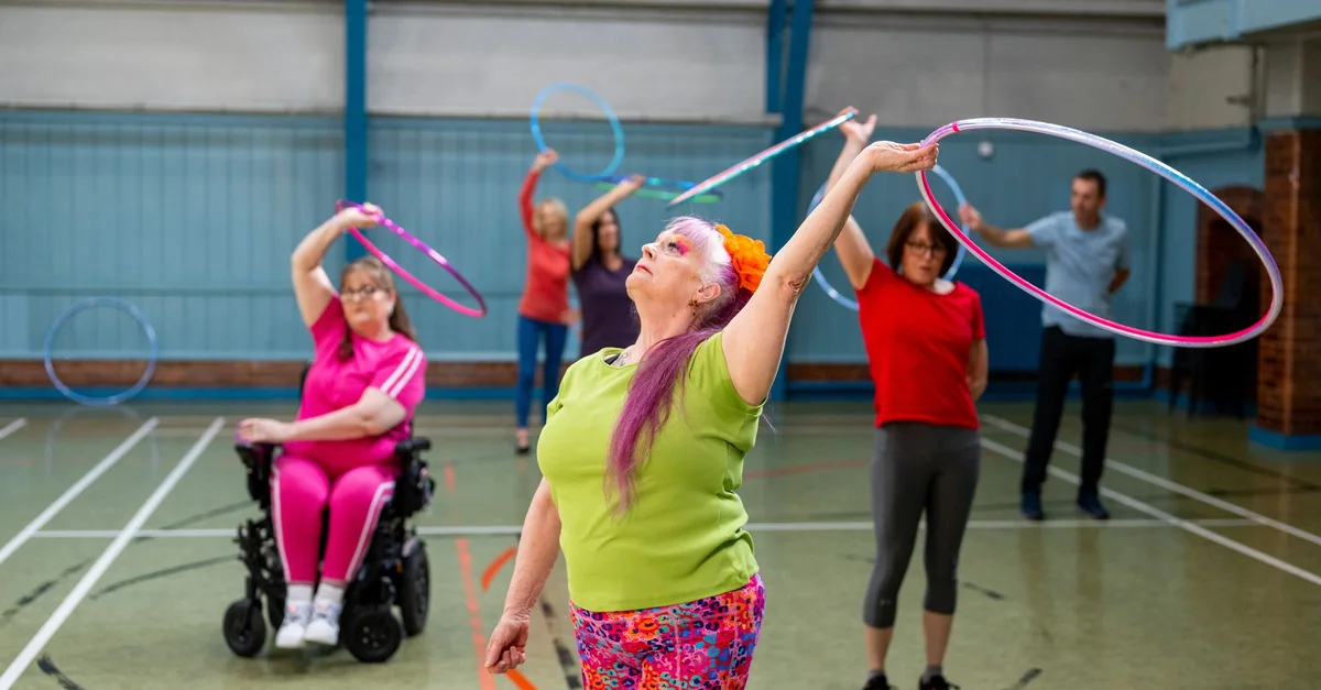 A group of adults, including seniors and a person in a wheelchair, enjoying a hula hoop exercise indoors.