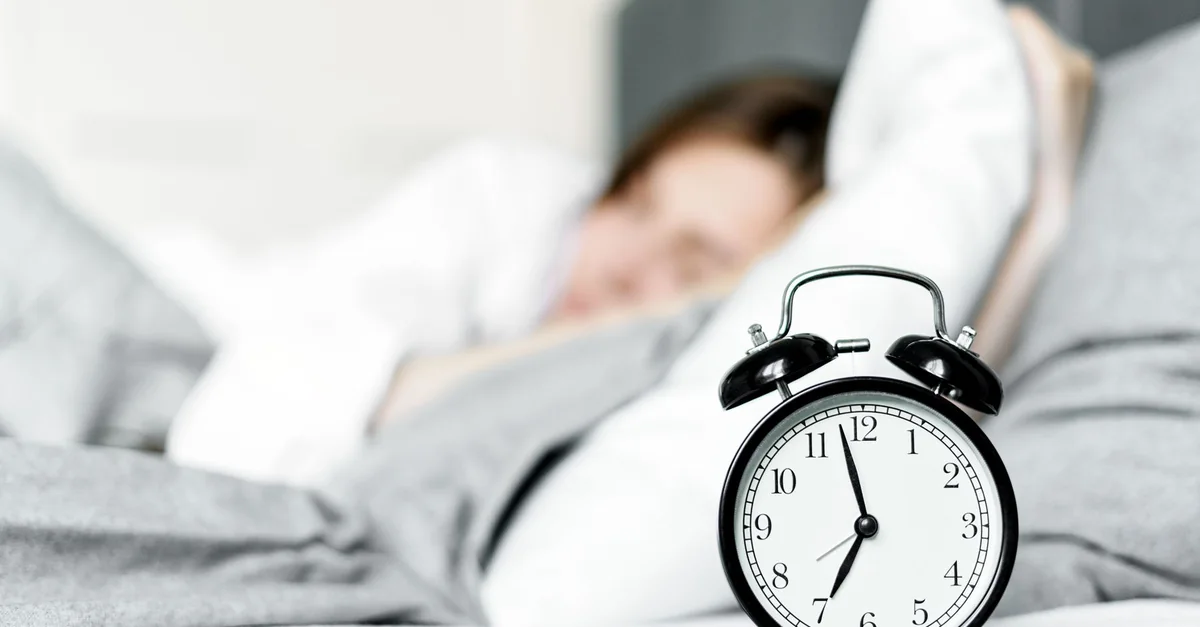 A retro alarm clock sits by a sleeping woman in a cozy bedroom setting.