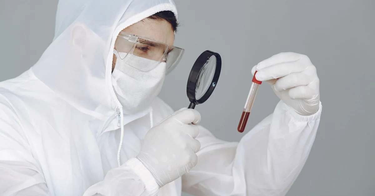 A scientist in protective gear examines a blood sample through a magnifying glass.