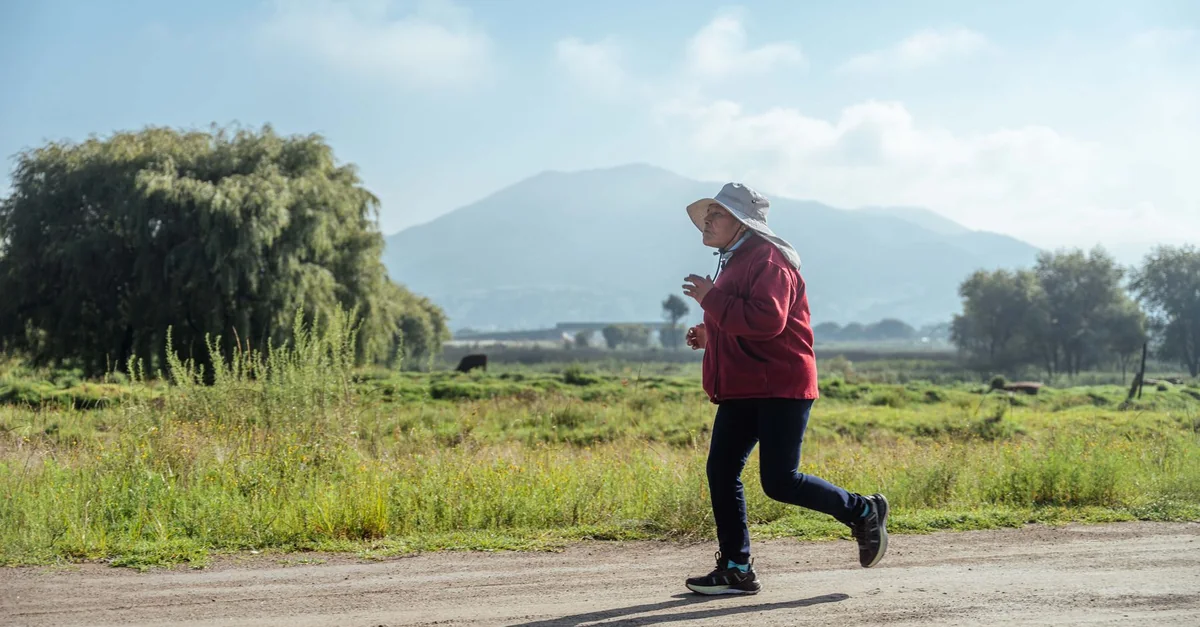 Senior woman jogging on a dirt road in a rural landscape with mountains in the background.