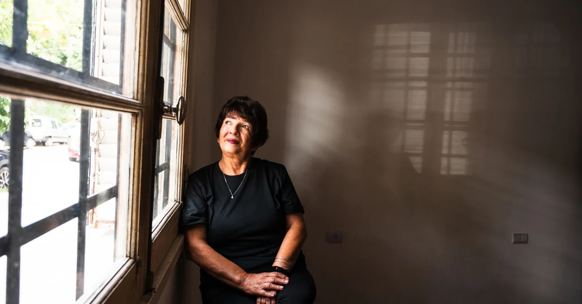 A thoughtful senior woman sitting by a window in a softly lit room, lost in contemplation.