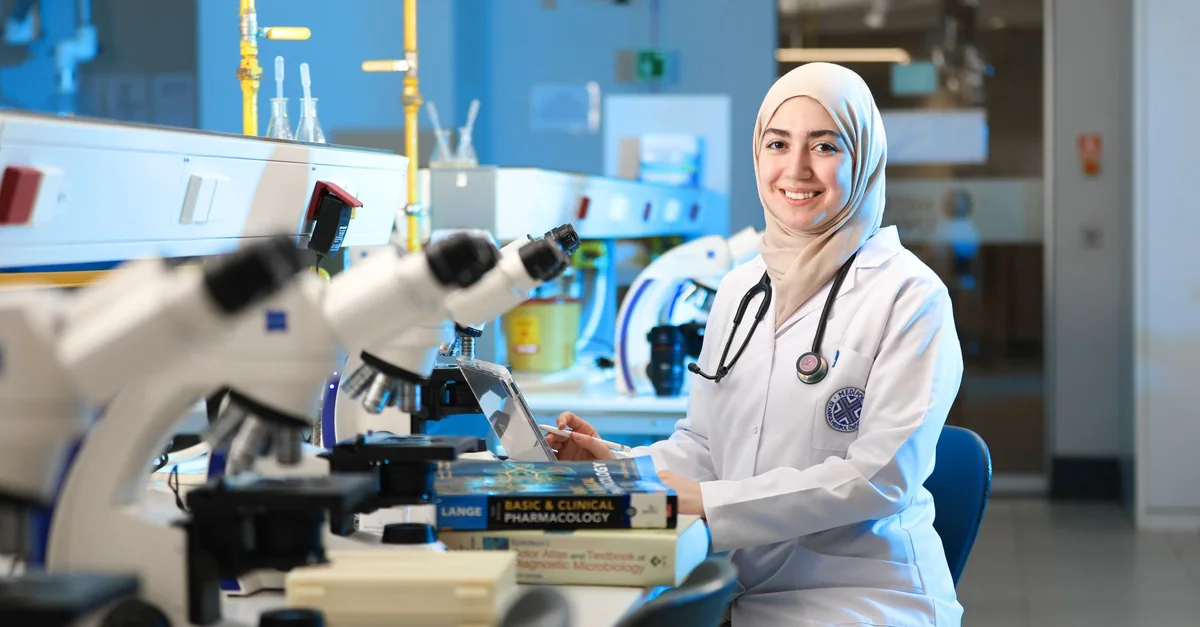 A confident female scientist in a lab coat with a hijab in a modern laboratory with microscopes and textbooks.