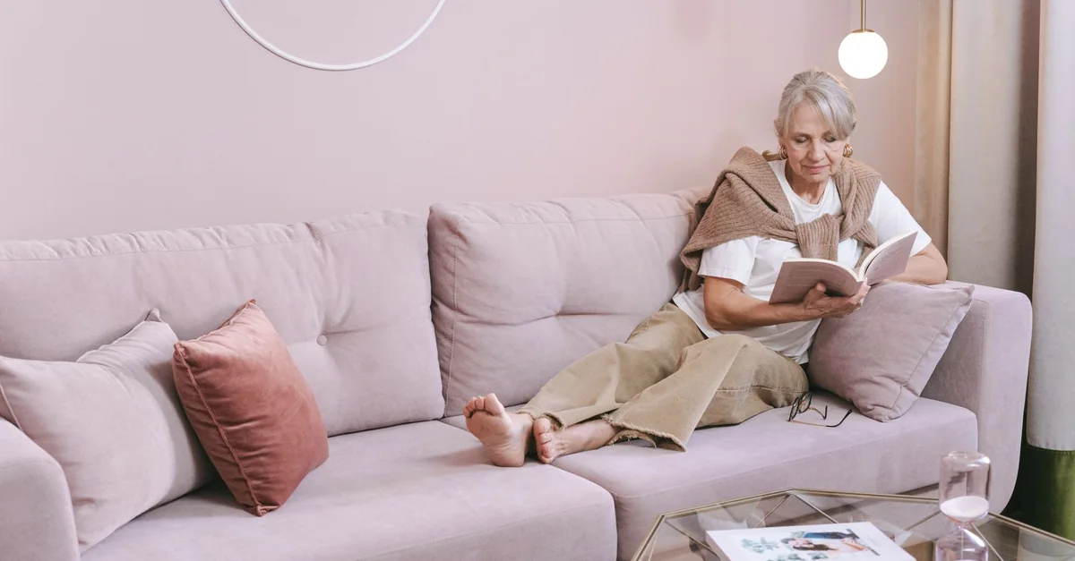 Senior woman relaxing and reading a book at home on a comfortable pink sofa.