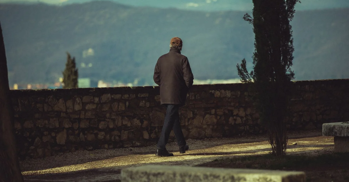 A lone senior adult walking along a stone path with a mountain backdrop under a clear sky.