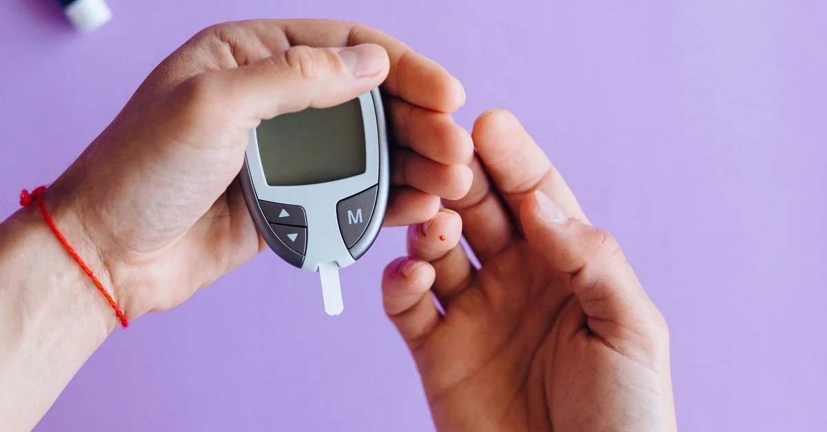 Hands holding a glucometer to test blood sugar levels on a purple background.